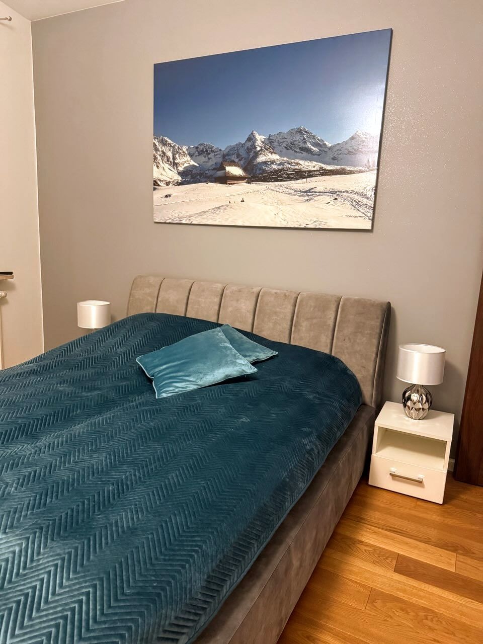 Beżowe łóżko tapicerowane z wezgłowiem w pionowe pasy, turkusowa narzuta w zygzaki i poduszki. Nad łóżkiem wisi obraz z górskim pejzażem.