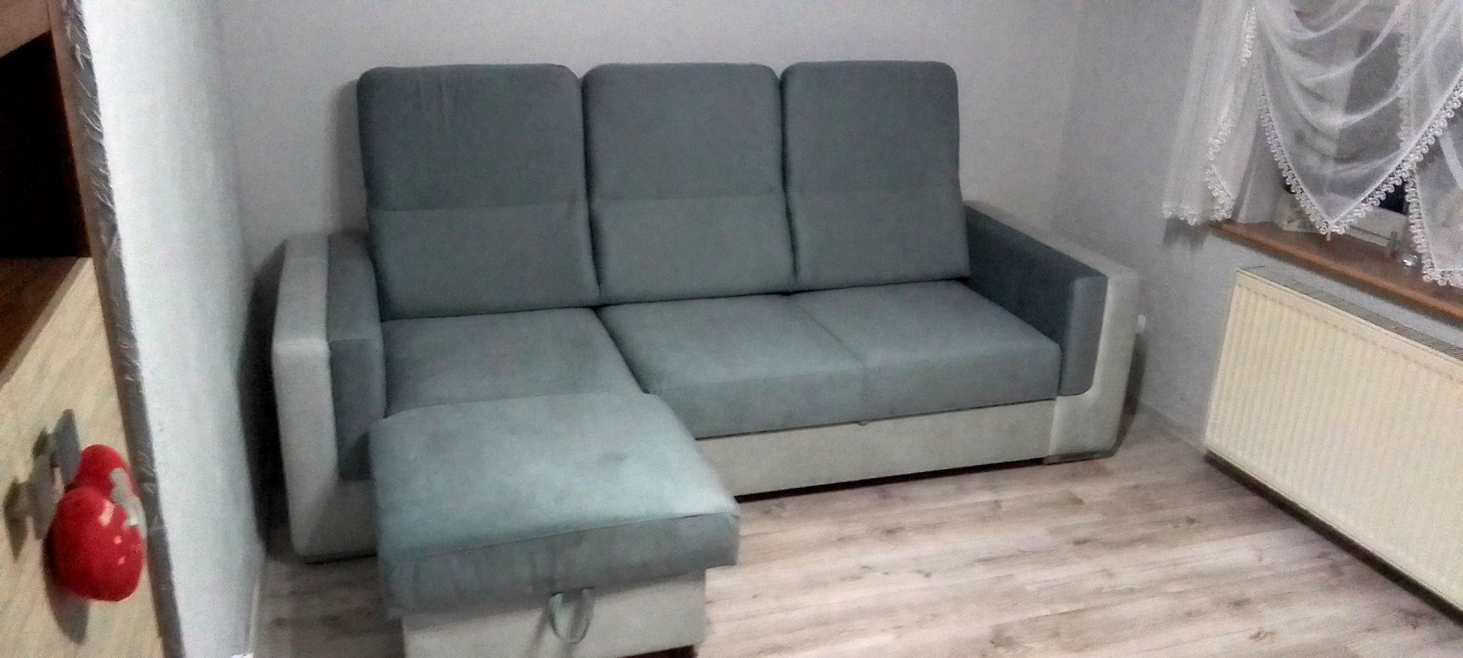 Szara, rozkładana sofa narożna z pufą w pokoju. Widoczny fragment okna z firanką i grzejnik. Ujęcie z niskiego kąta, podkreślające przestronność mebla.