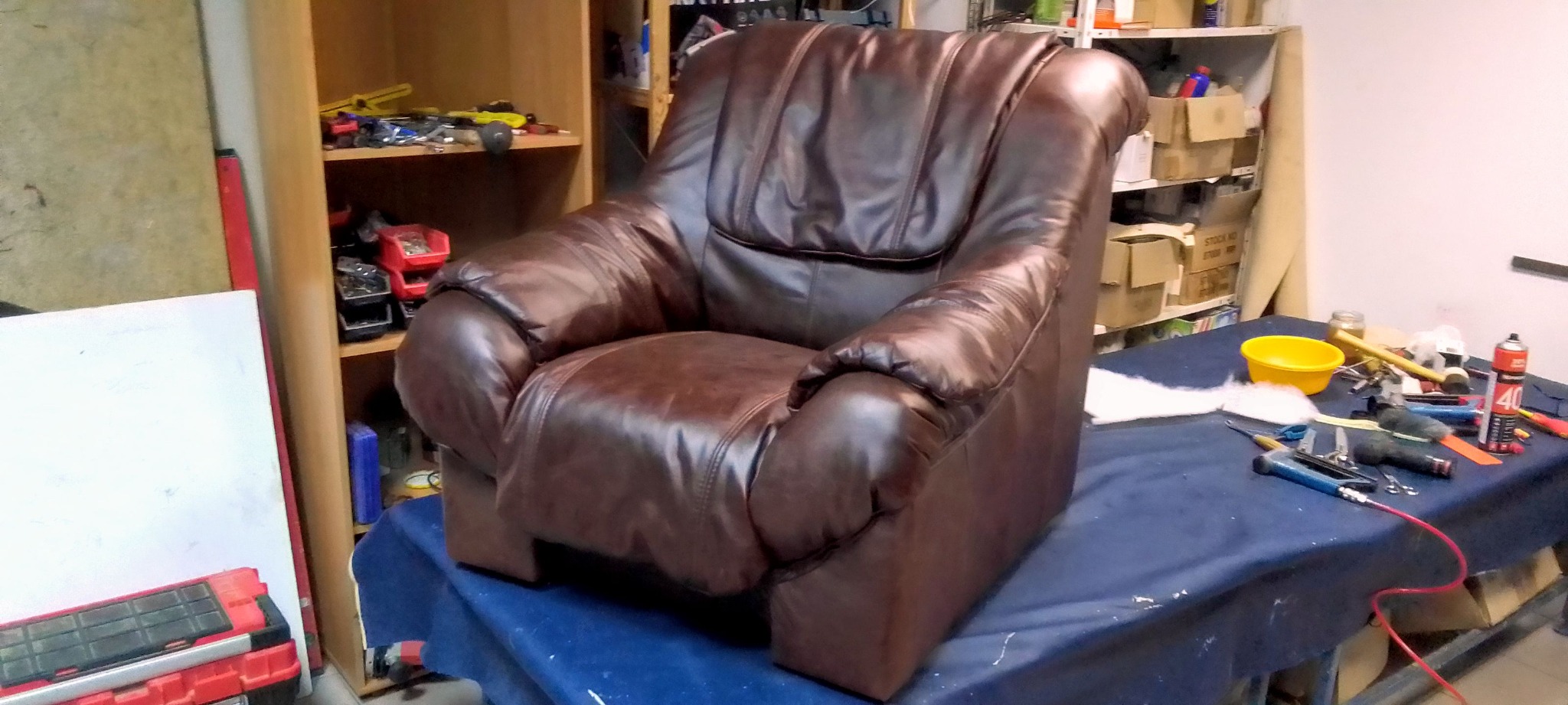 Brązowy, skórzany fotel po renowacji tapicerskiej, ustawiony w warsztacie z narzędziami i materiałami tapicerskimi na stole. Widoczne zszywacze, wata i inne akcesoria.