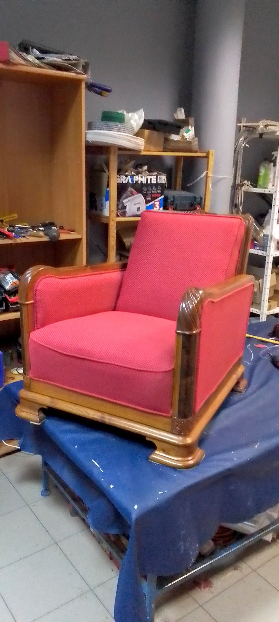 Odrestaurowany fotel w stylu art deco z czerwoną tapicerką i drewnianymi elementami, stojący na stole w warsztacie tapicerskim. Widoczne narzędzia i materiały.