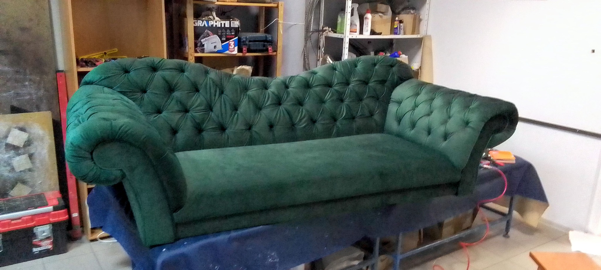 Szmaragdowa leżanka Chesterfield z głębokim pikowaniem, ustawiona w warsztacie tapicerskim, czeka na wykończenie. Widoczne narzędzia i materiały w tle.