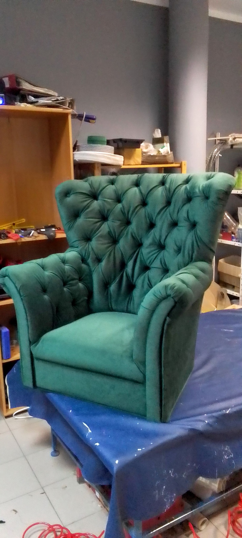 Szmaragdowy fotel typu chesterfield z głębokim pikowaniem, ustawiony na niebieskiej folii ochronnej w warsztacie tapicerskim. Widoczne narzędzia i materiały w tle.
