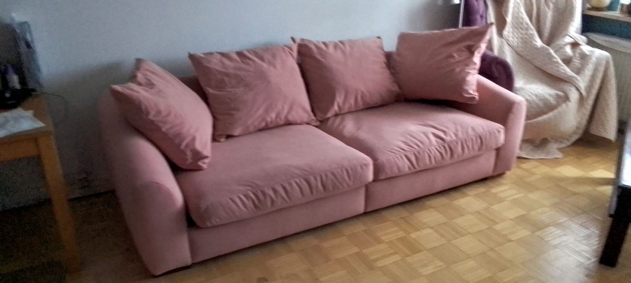 Różowa, dwuosobowa sofa z poduszkami w salonie na parkiecie. W tle widoczny fotel przykryty beżowym pledem. Sofa o miękkich liniach, w stylu skandynawskim.