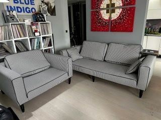 Szary komplet wypoczynkowy: sofa i fotel w geometryczny wzór, z czarną lamówką, na tle regału z płytami i abstrakcyjnego obrazu.