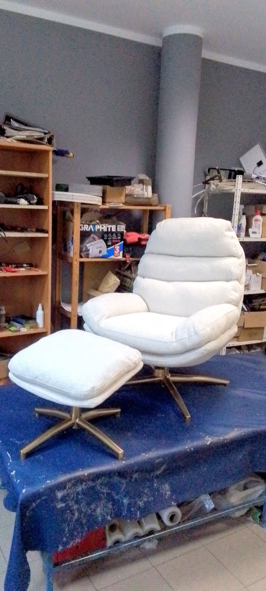 Biały, tapicerowany fotel z podnóżkiem na złotych nogach, sfotografowany w warsztacie tapicerskim. Widoczne półki z narzędziami i materiałami.