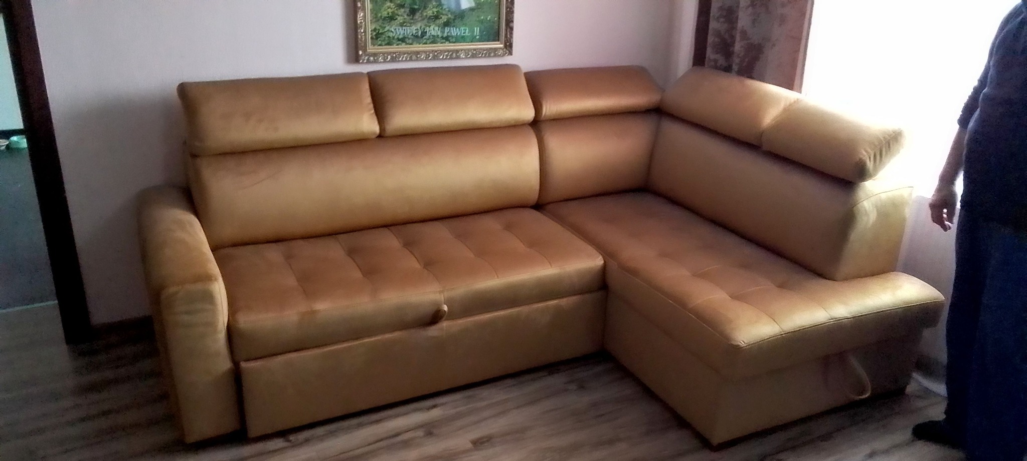 Złoty narożnik z funkcją spania i pojemnikiem na pościel, widoczny fragment osoby. Tapicerka z przeszyciami. Nad sofą obraz w złotej ramie.