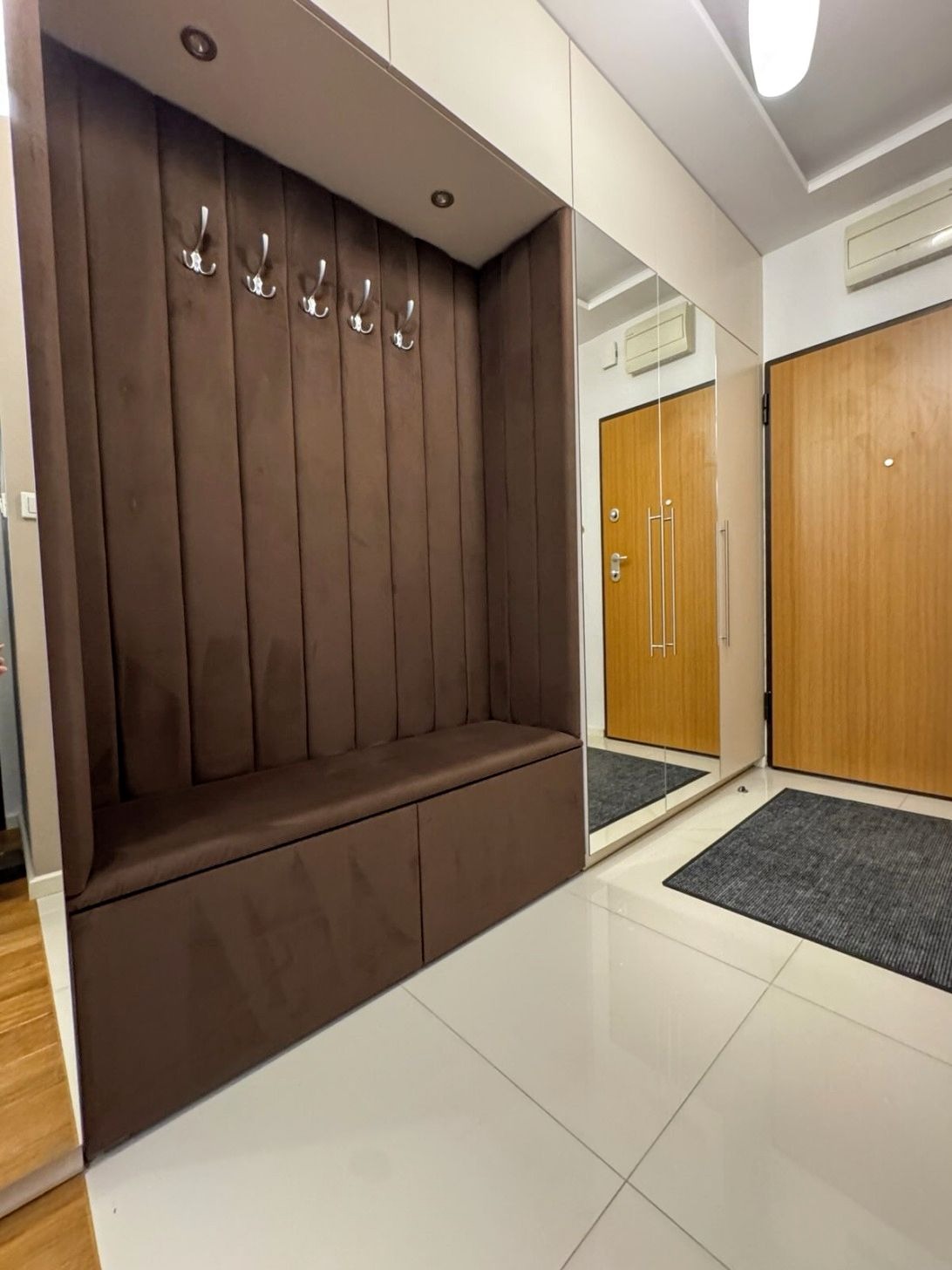 Tapicerowany, brązowy przedpokój z siedziskiem i wieszakami, wbudowany w szafę z lustrem. Nowoczesny design, jasne płytki podłogowe.