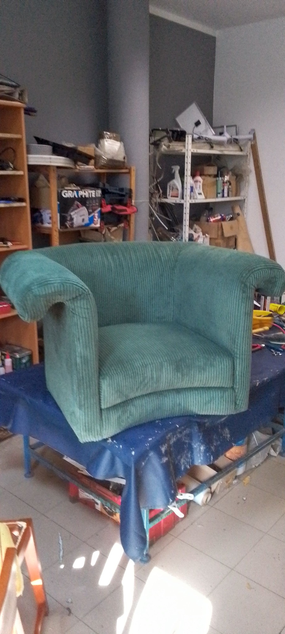 Fotel w zielonej, prążkowanej tapicerce, stojący na stole przykrytym niebieską tkaniną. W tle regały z narzędziami i materiałami.