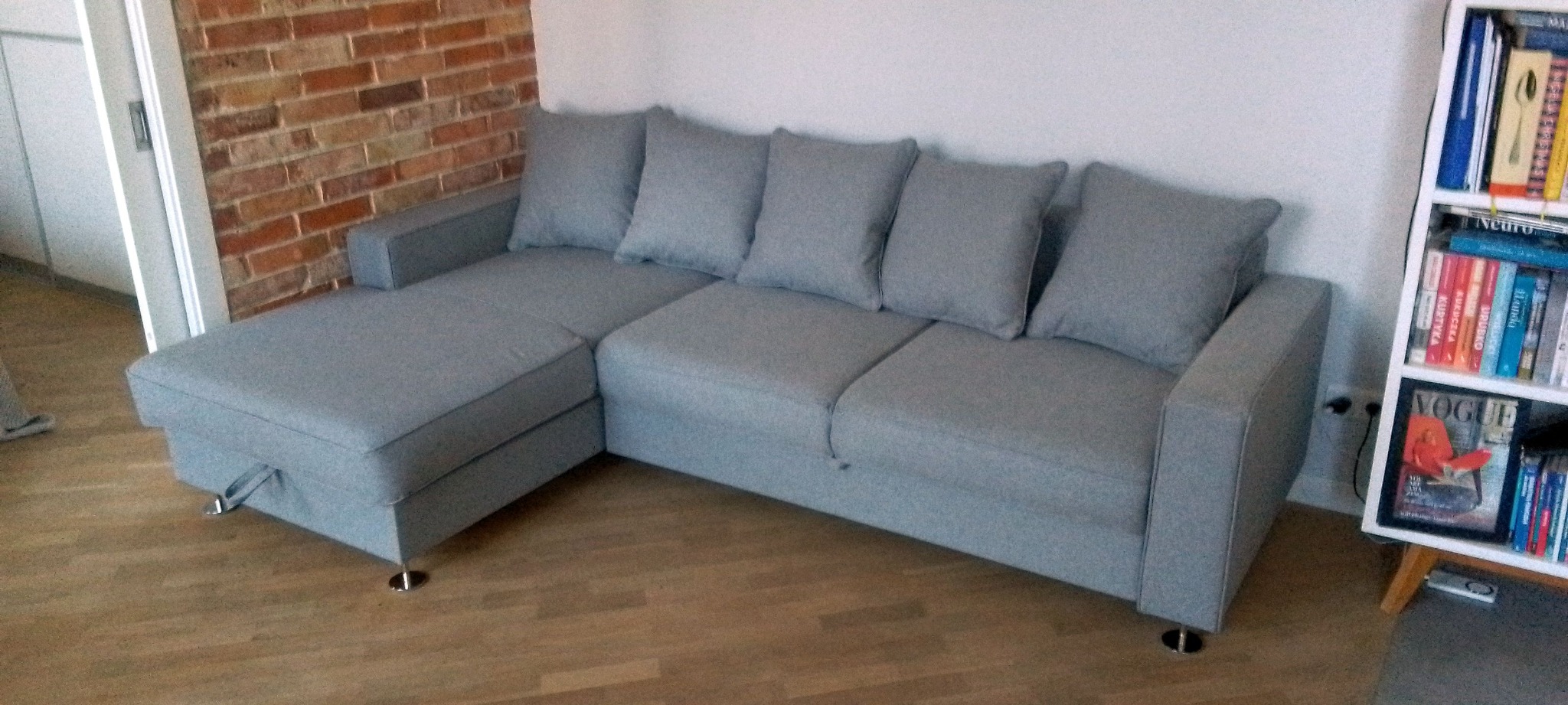 Szara sofa narożna z poduszkami, ustawiona w salonie z ceglaną ścianą i regałem na książki. Realizacja z Dobrego Miasta. Minimalistyczny design.