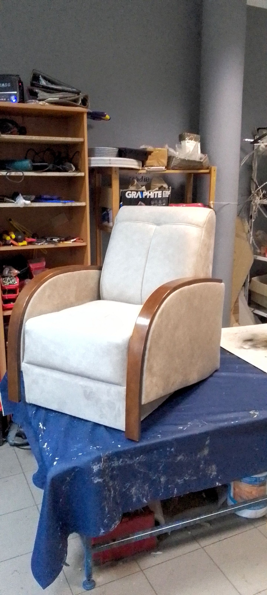 Kremowy fotel z drewnianymi podłokietnikami, stojący na stole w warsztacie tapicerskim. W tle półki z narzędziami i materiałami.