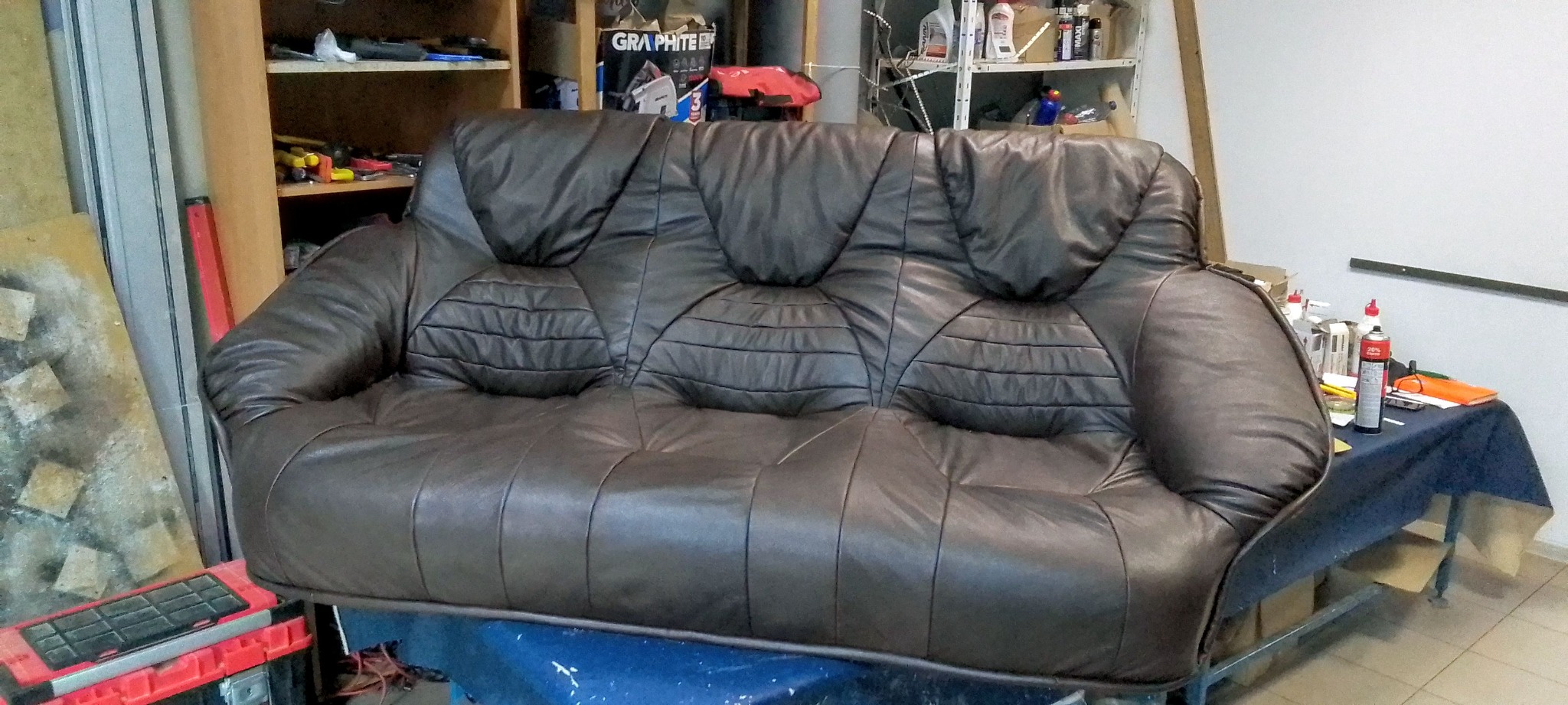 Brązowa, skórzana sofa z trzema siedziskami, z charakterystycznym pikowaniem na oparciach, stoi w warsztacie tapicerskim. Widoczne narzędzia i materiały.