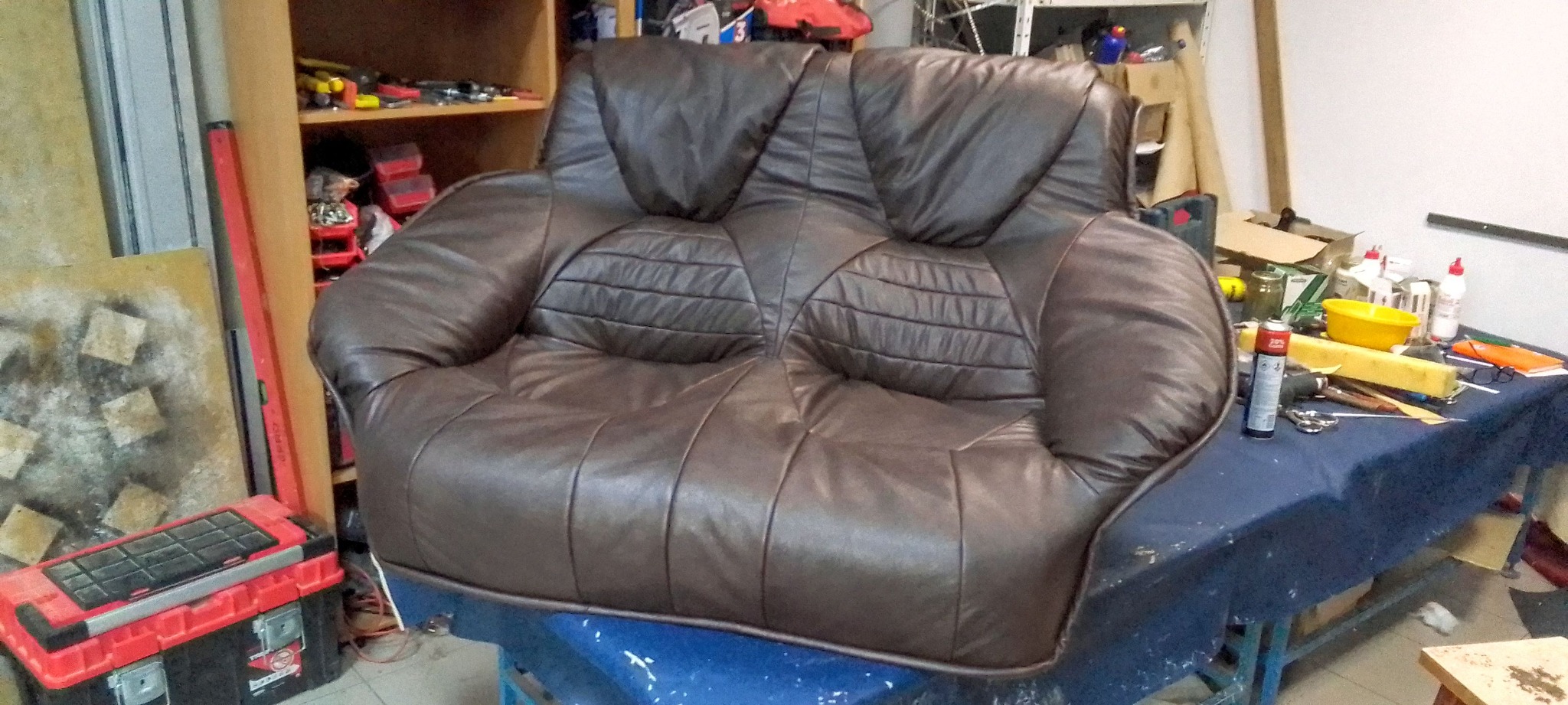 Brązowy, skórzany fotel dwuosobowy w warsztacie tapicerskim. Widoczne narzędzia i materiały, co sugeruje renowację lub produkcję. Charakterystyczne przeszycia na siedzisku.