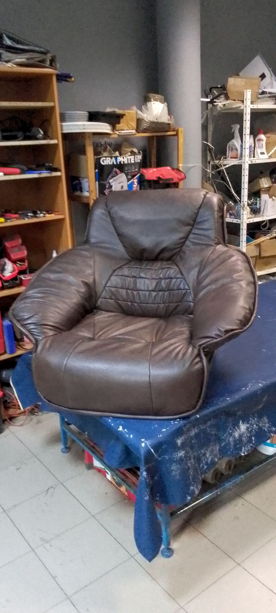 Brązowy, skórzany fotel klubowy po renowacji tapicerskiej, ustawiony na stole w warsztacie. Widoczne detale pikowania i przeszycia. W tle regały z narzędziami.