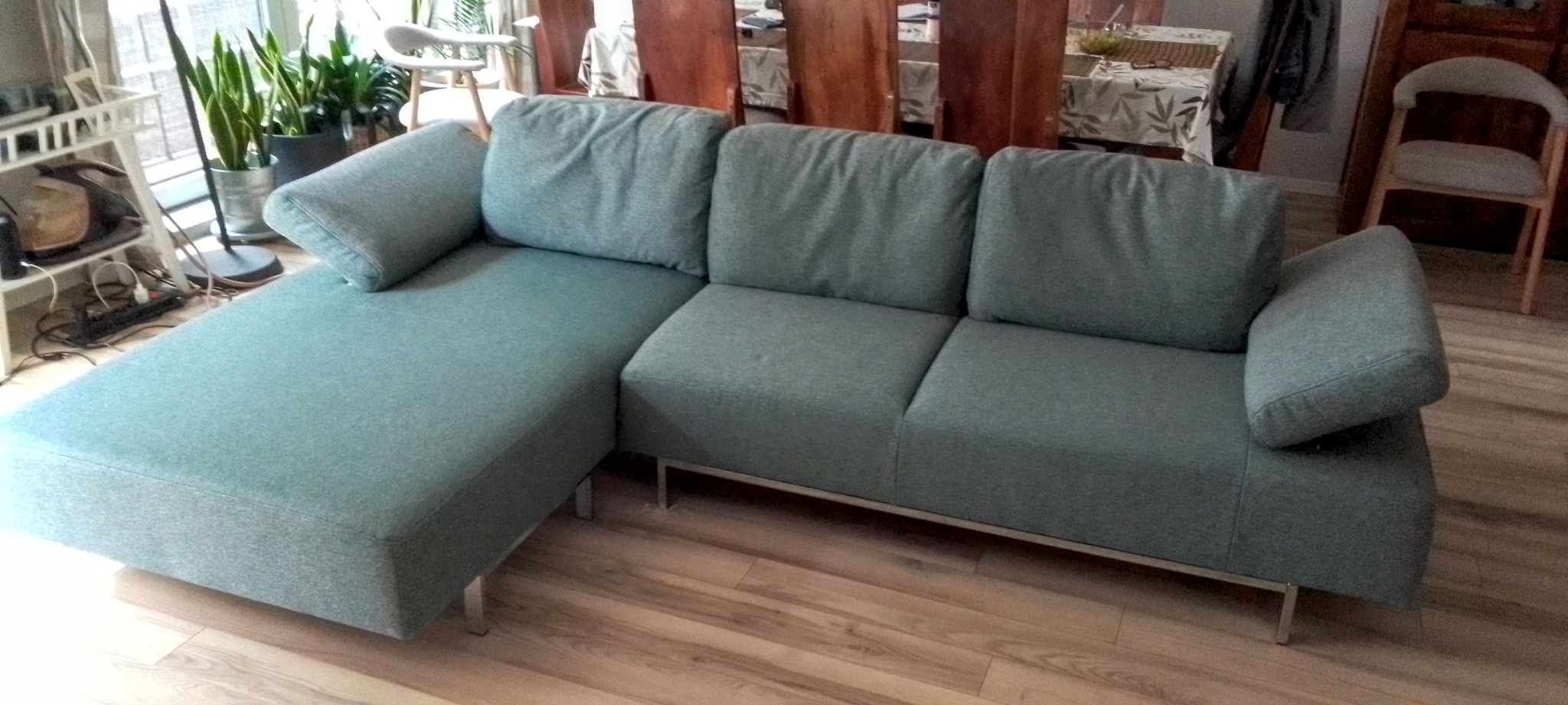 Turkusowa sofa z szezlongiem na metalowych nóżkach, widok z przodu, na drewnianej podłodze. W tle stół i krzesła, doniczki z roślinami.