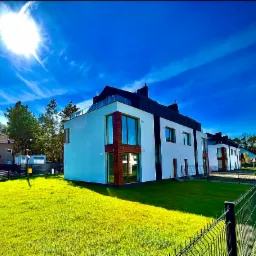 Nowoczesne, szeregowe domy z białymi elewacjami i drewnianymi akcentami, widoczne spod niskiego kąta na tle intensywnie niebieskiego nieba z promieniami słońca i zielonego trawnika z metalowym...