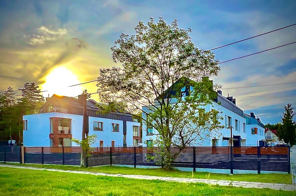 Nowoczesne, jasnoniebieskie domy szeregowe z ciemnymi dachami i drewnianymi akcentami, ogrodzone poziomym, ciemnym płotem, widoczne na tle zachodzącego słońca i błękitnego nieba z chmurami.