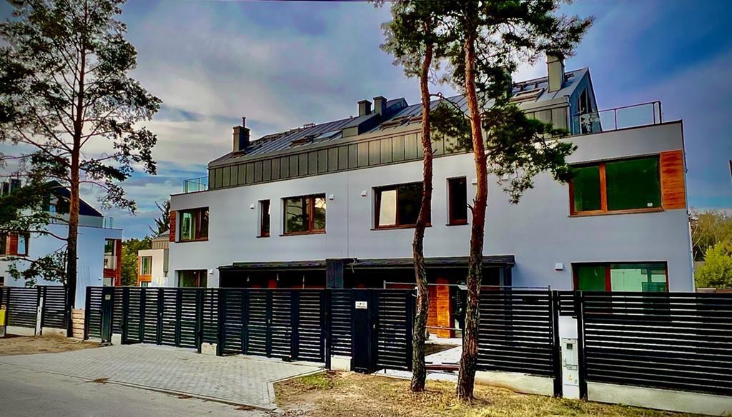 Nowoczesny, dwukondygnacyjny dom z płaskim dachem i czarnym ogrodzeniem panelowym, widoczny z poziomu chodnika, z drzewami po bokach kadru.