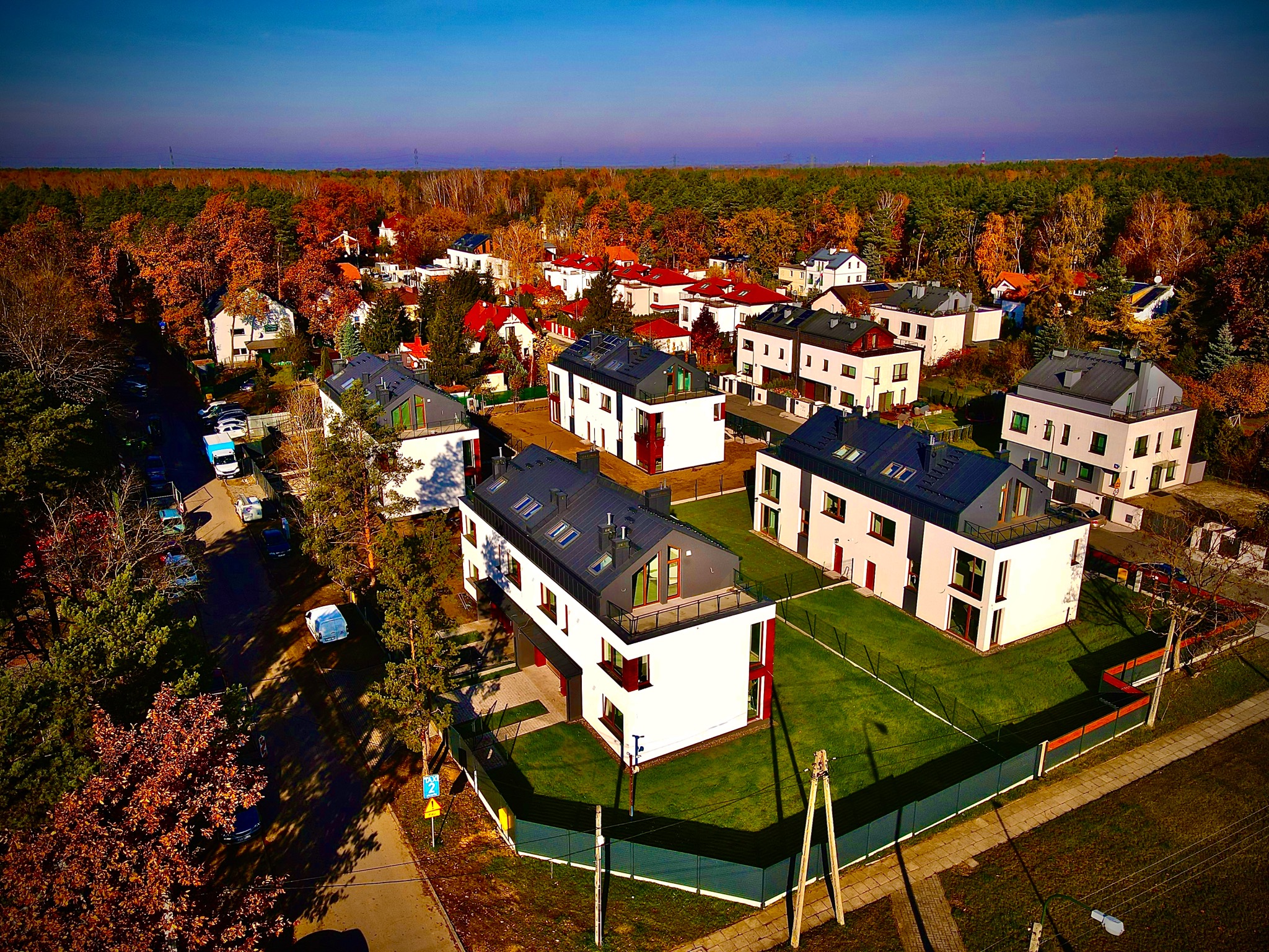 Osiedle nowoczesnych, dwukondygnacyjnych domów jednorodzinnych z płaskimi dachami i dużymi oknami, widziane z lotu ptaka w jesiennej scenerii, z lasem w tle.