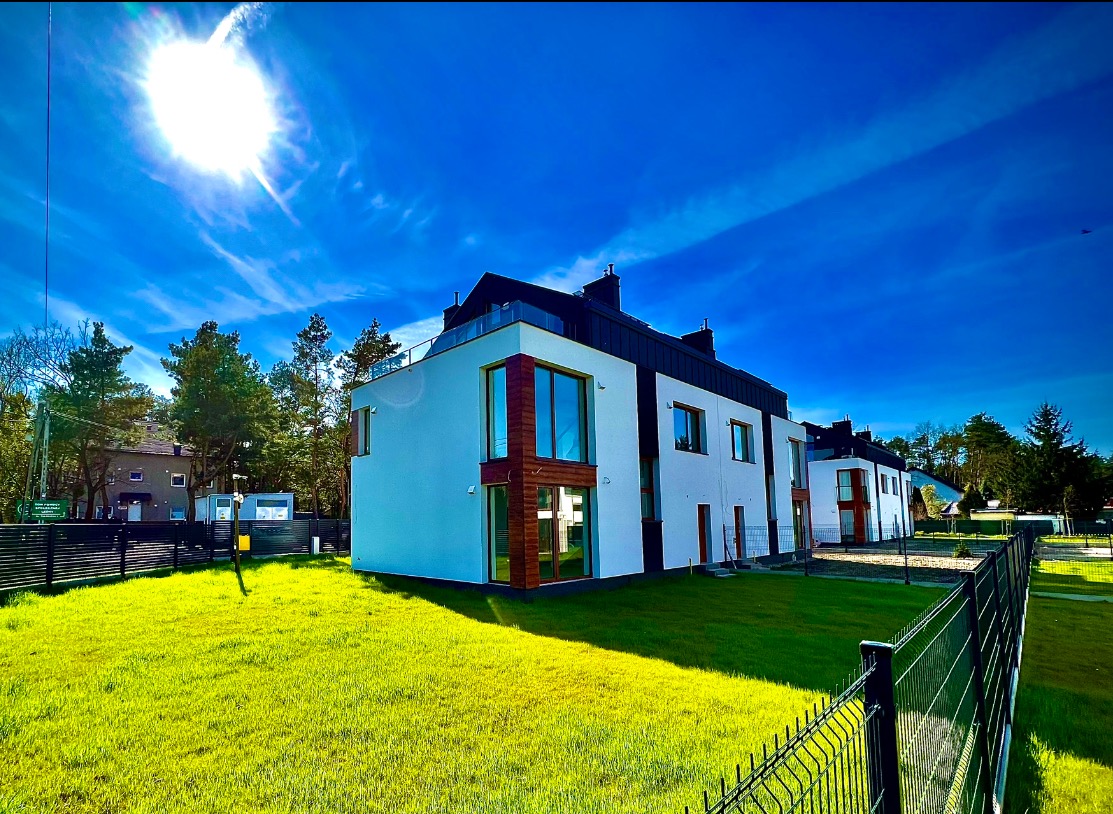 Nowoczesne, szeregowe domy z białymi elewacjami i drewnianymi akcentami, widoczne spod niskiego kąta na tle intensywnie niebieskiego nieba z promieniami słońca i zielonego trawnika z metalowym...