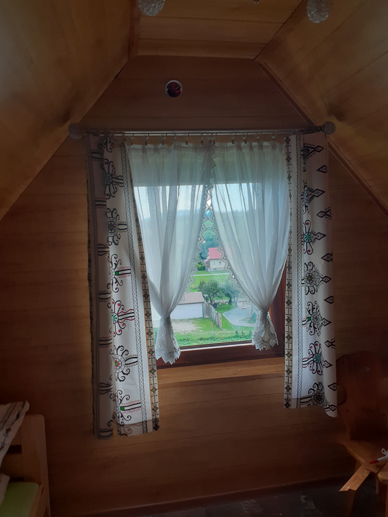 Okno w drewnianym pokoju na poddaszu z białymi, wzorzystymi zasłonami, przez które widać zielony krajobraz