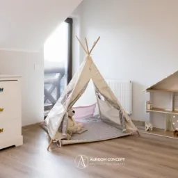 Pokój dziecięcy na poddaszu: biała komoda z motylami, tipi z pluszowym misiem w środku, drewniany domek dla lalek z oświetleniem LED.