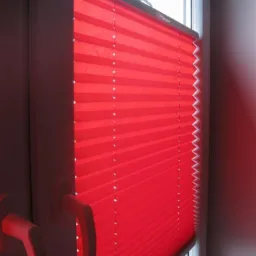 Czerwona, plisowana roleta okienna zamontowana w oknie PCV z widocznym uchwytem do regulacji, data na szybie: 01/02/2013.