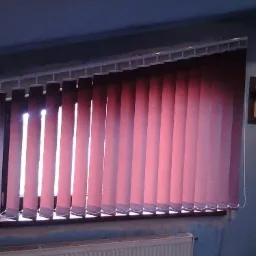 Czerwono-brązowe pionowe panele okienne z widocznym mechanizmem mocowania na tle okna z delikatnym prześwitem światła dziennego i fragmentem grzejnika pod parapetem.