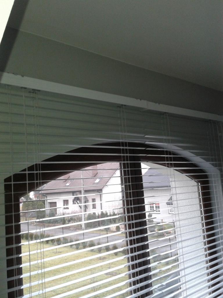 Wnętrze z oknem o łukowatym kształcie, przesłoniętym poziomymi żaluzjami w kolorze białym, z widokiem na dom i ogród.