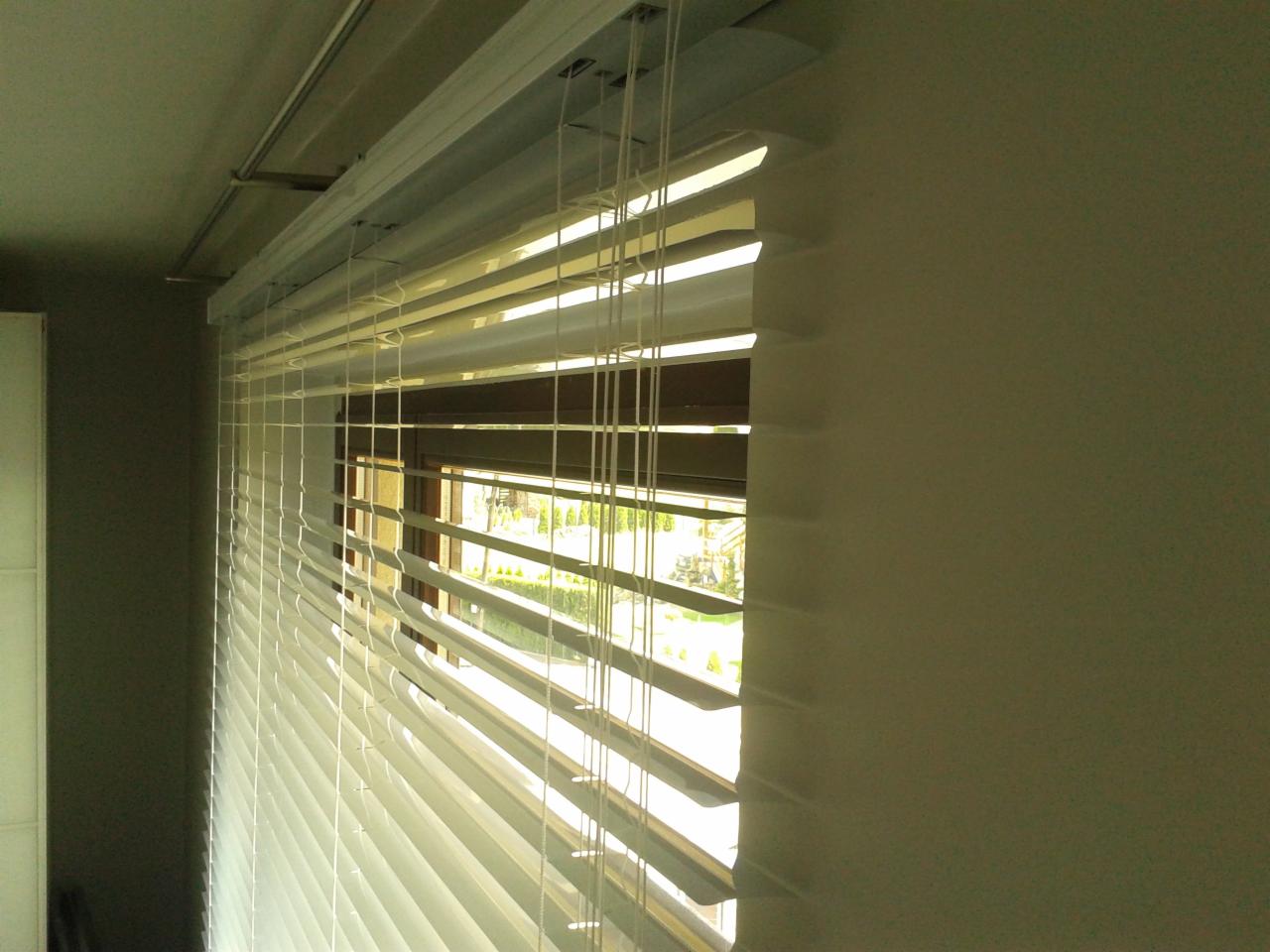 Zamknięte, białe poziome osłony okienne z widocznym mechanizmem sznurkowym i fragmentem ramy okiennej z widokiem na zieleń za oknem.