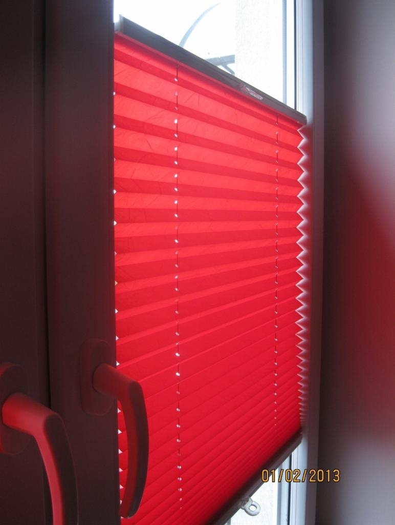 Czerwona, plisowana roleta okienna zamontowana w oknie PCV z widocznym uchwytem do regulacji, data na szybie: 01/02/2013.