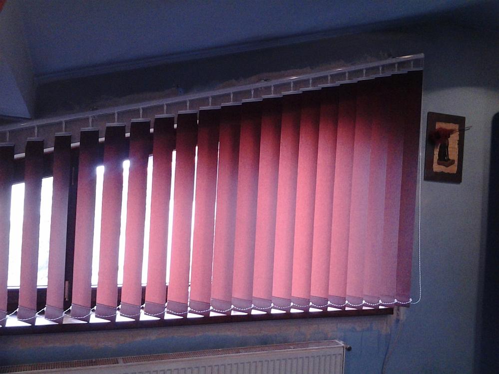 Czerwono-brązowe pionowe panele okienne z widocznym mechanizmem mocowania na tle okna z delikatnym prześwitem światła dziennego i fragmentem grzejnika pod parapetem.