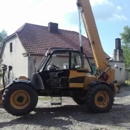 Ż&oacute;łto-czarna maszyna z wysięgnikiem na piaszczystym podłożu przed budynkiem z czerwonym dachem.