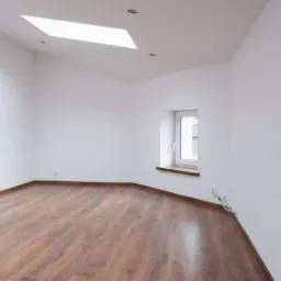 Puste, jasne pomieszczenie z drewnianą podłogą, białymi ścianami, oknem i świetlikiem w suficie, przygotowane do wykończenia.