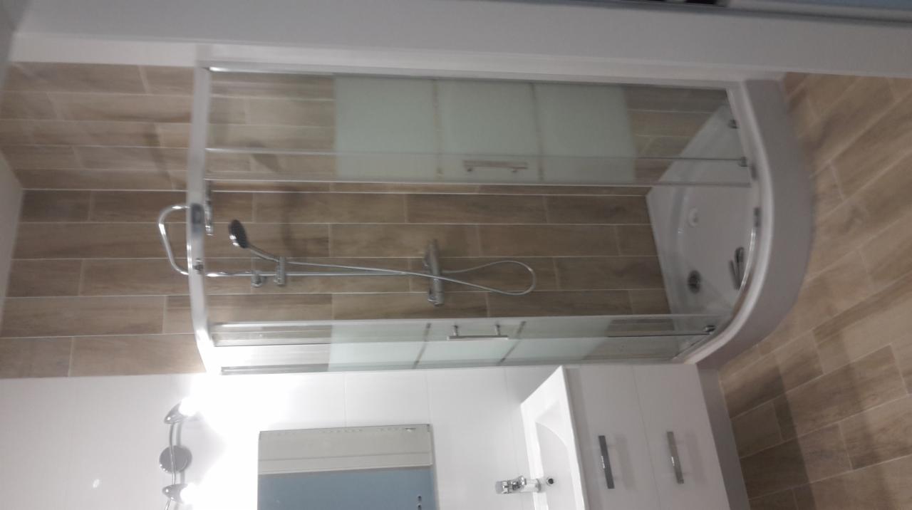 Nowoczesna kabina prysznicowa z hydromasażem, obudowana płytkami imitującymi drewno, z widoczną umywalką z szafką i lustrem w małej łazience.