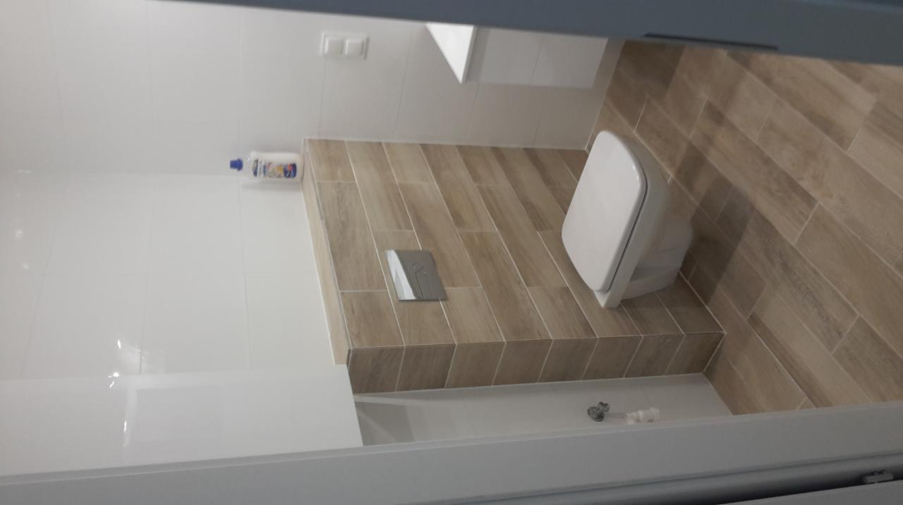 Nowoczesna toaleta z podwieszaną muszlą, obudowana płytkami imitującymi drewno, z widocznym przyciskiem spłukującym i butelką środka czyszczącego na półce.