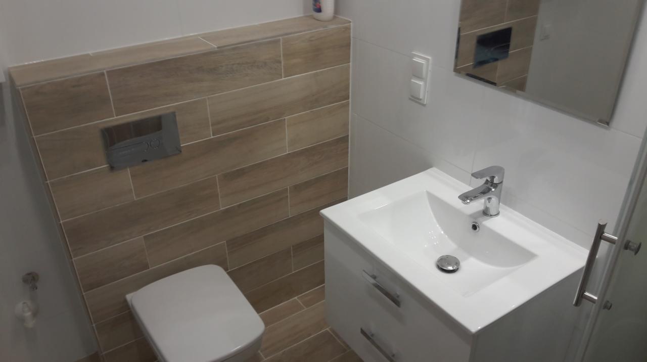 Nowoczesna łazienka z beżowymi płytkami imitującymi drewno, białą umywalką z szafką i wiszącą toaletą, ukazana z perspektywy lekko z góry.