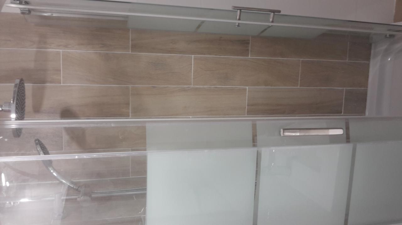 Wykończona kabina prysznicowa z przesuwnymi, matowymi drzwiami, zamontowana na tle ściany wyłożonej podłużnymi, drewnopodobnymi płytkami ceramicznymi. Widoczna chromowana deszczownica i słuchawka...