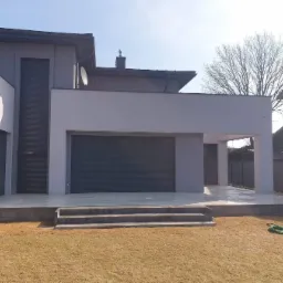 Nowoczesny dom z szarą elewacją i tarasem wyłożonym jasnymi płytami, schody z ciemnoszarego kamienia prowadzą na trawnik, widoczny zwinięty wąż ogrodowy.