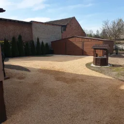 Wykończony teren posesji z wysypanym żwirem, widoczny garaż blaszany, studnia zadaszona, budynki gospodarcze, młode tuje i błękitne niebo.