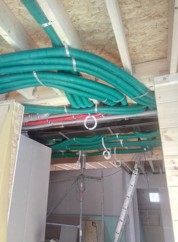 Zielone, karbowane rury wentylacyjne przymocowane do drewnianego stropu za pomocą metalowych obejm; widoczne również rury czerwone i szare oraz fragment ściany z płyty gipsowej i drabina.