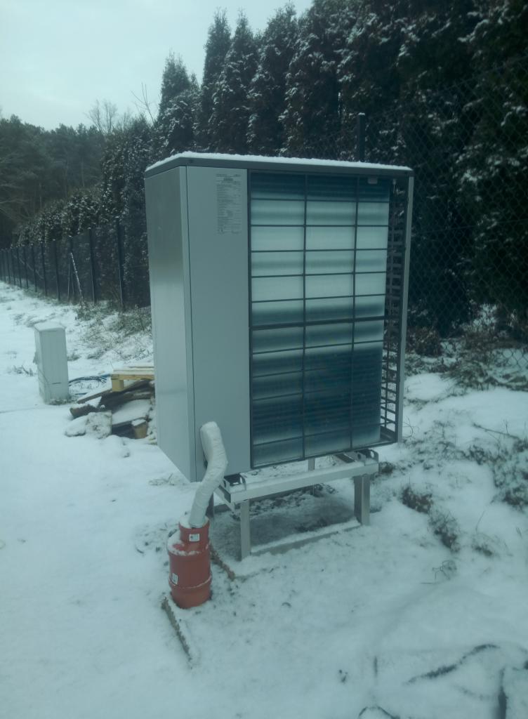 Zewnętrzna jednostka pompy ciepła, zamontowana na metalowej konstrukcji, z podłączonym izolowanym przewodem odprowadzającym skropliny do pomarańczowego zbiornika, na tle ośnieżonego ogrodu.