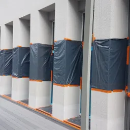 Przygotowanie elewacji budynku usługowego do malowania: zabezpieczone folią i taśmą okna oraz kolumny, widoczny fragment elewacji z fakturą i fragment przechodzącego mężczyzny.