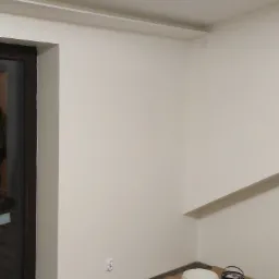 Świeżo pomalowany narożnik pokoju z widocznymi puszkami farb Admiral i Sigma oraz akcesoriami malarskimi na kartonowym zabezpieczeniu podłogi.