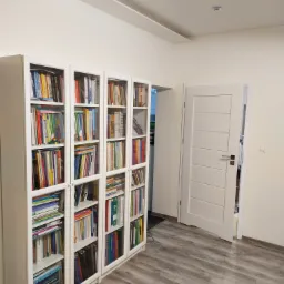 Białe regały z przeszklonymi drzwiczkami wypełnione książkami i segregatorami, uzupełnione drewnopodobną podłogą i białą ścianą z drzwiami.