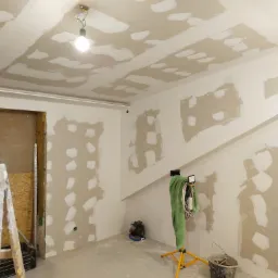 Pokój w trakcie prac przygotowawczych do malowania: ściany i sufit z widocznymi łatami gipsowymi, drabina, lampa na statywie z ręcznikiem i wiadro z narzędziami.