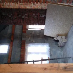 Surowe wnętrze poddasza w trakcie adaptacji: widoczne cegły, bloczki, drewniane belki, płyta OSB oparta o ścianę, pakiet desek, butelka wody i drabina.