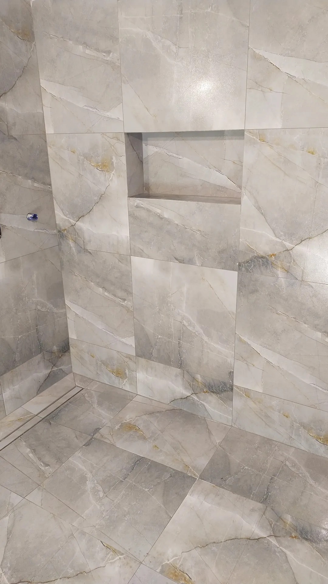 Wykończona łazienka z szarymi płytkami imitującymi marmur i wnęką w ścianie, widoczny odpływ liniowy w podłodze.