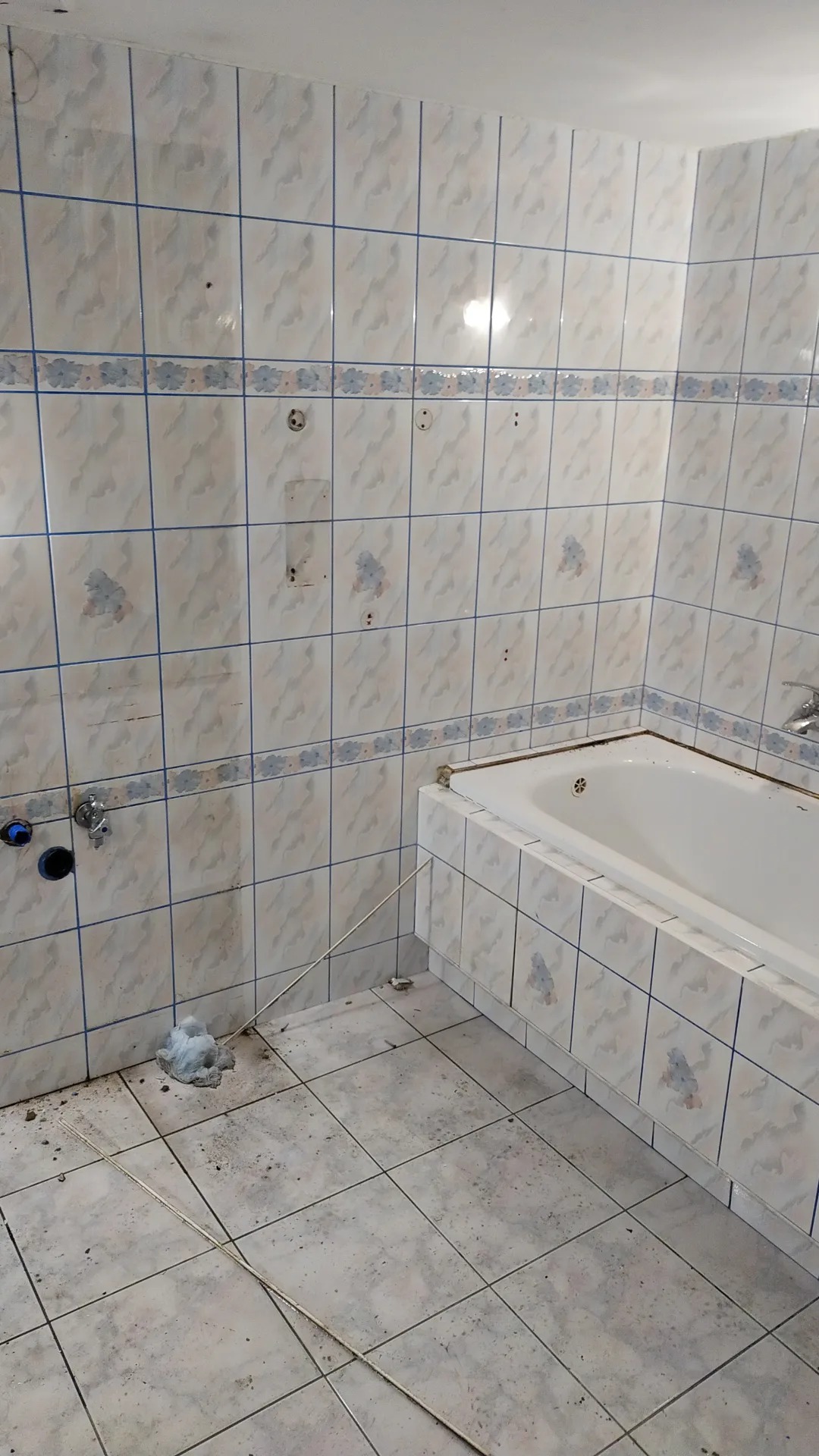 Łazienka w trakcie demontażu płytek, widoczne ślady po usuniętych płytkach na ścianie, stara wanna obłożona płytkami, podłoga z płytek w trakcie czyszczenia, instalacja wodna bez baterii.