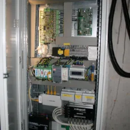 Otwarta szafa sterownicza z widocznymi płytkami drukowanymi, przewodami, zasilaczem awaryjnym, modułami sterującymi i elementami zabezpieczeń, zamontowana na szarej ścianie.