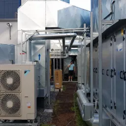 Instalacja wentylacyjna na dachu budynku, widoczny agregat Daikin, rury wentylacyjne, centrala wentylacyjna, pracownik w tle rozmawia przez telefon.